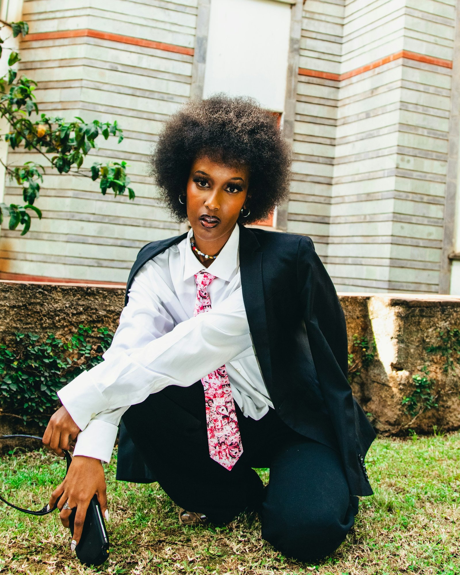 Photo by Dwayne joe on Unsplash of a melanated woman with an afro is wearing a dark-colored suit, white dress shirt, and bright multi-colored tie. In the right hand, there appears to be a phone or similar-shaped device. The left hand, with what appears to be sunglasses, crosses the right hand. The woman is partially kneeling on the short green grass. The woman also wears earrings and a necklace.