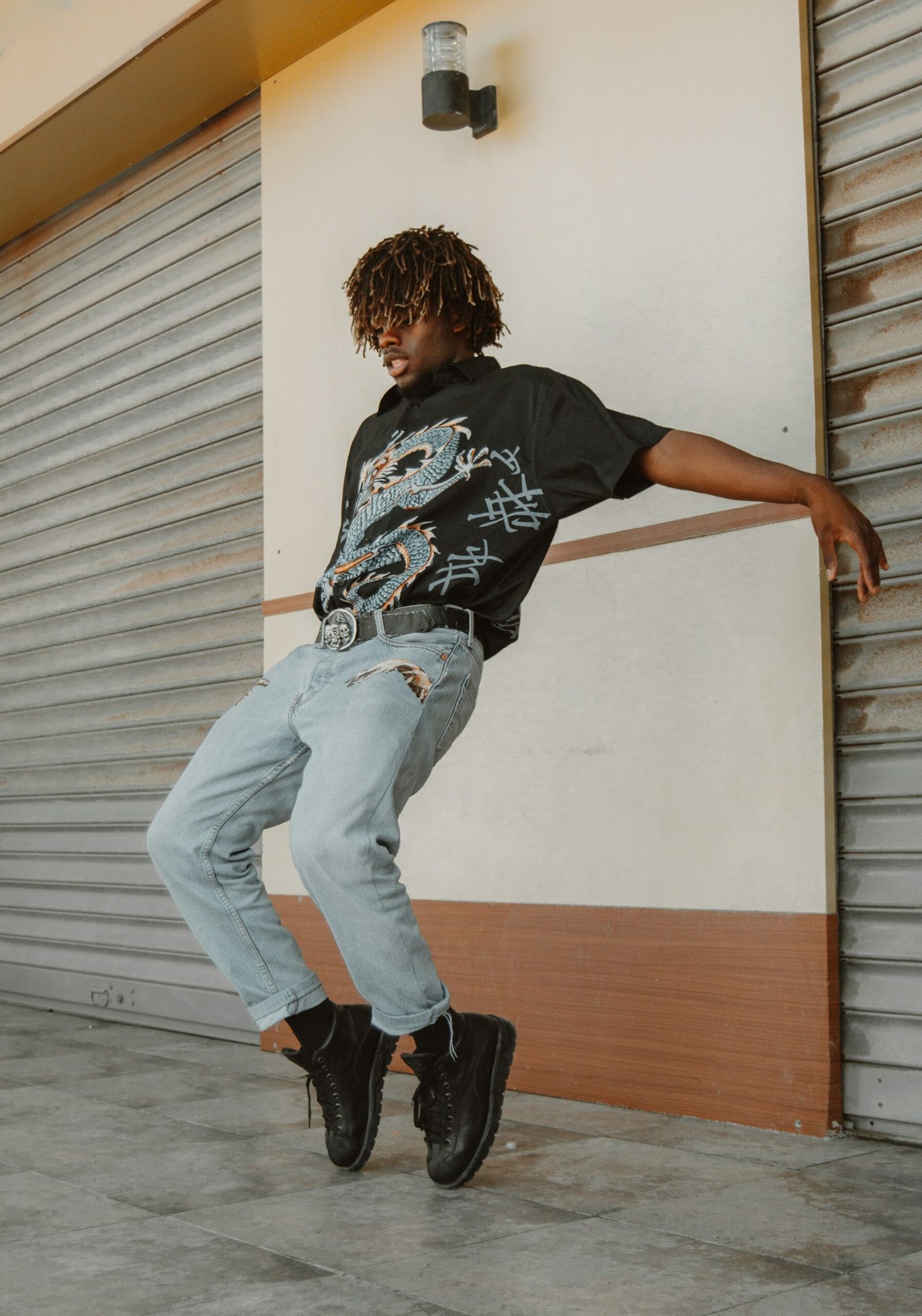Photo by Houcine Ncib on Unsplash. A young person with dark & light-colored locs wears a t-shirt, jeans and dark-colored shoes. The person pushes their arms back, while balancing on the toes of their shoes. Behind the model appears to be grey roller shutters. Photo details read: Photo of a male model dancing and celebrating life and youth.